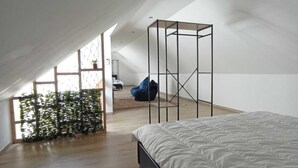 Une chambre spacieuse située dans les combles, avec un grand lit, un coin salon avec un fauteuil et une vue sur l’extérieur par une fenêtre.