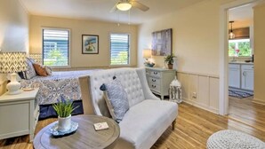 Un salon spacieux avec un grand canapé, une table à manger et un ventilateur de plafond. Il y a deux fenêtres équipées de stores, un coin cuisine à l’arrière-plan et une plante décorative sur la table.