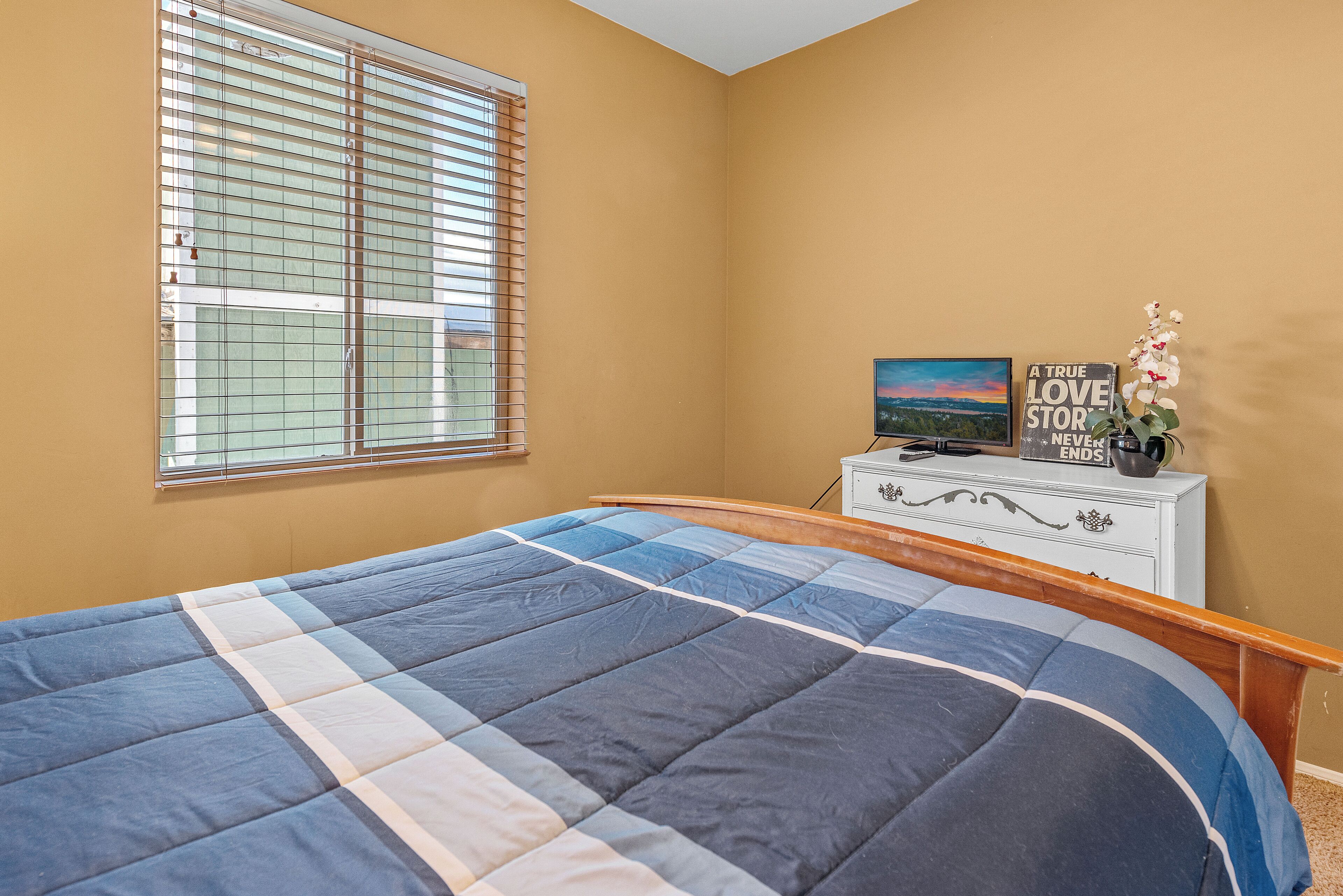 Een slaapkamer met een bed, een dressoir, een raam met zonwering en een decoratief object op het dressoir.