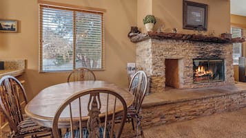 A stone fireplace with a fire, a dining table with chairs, and a window with blinds.