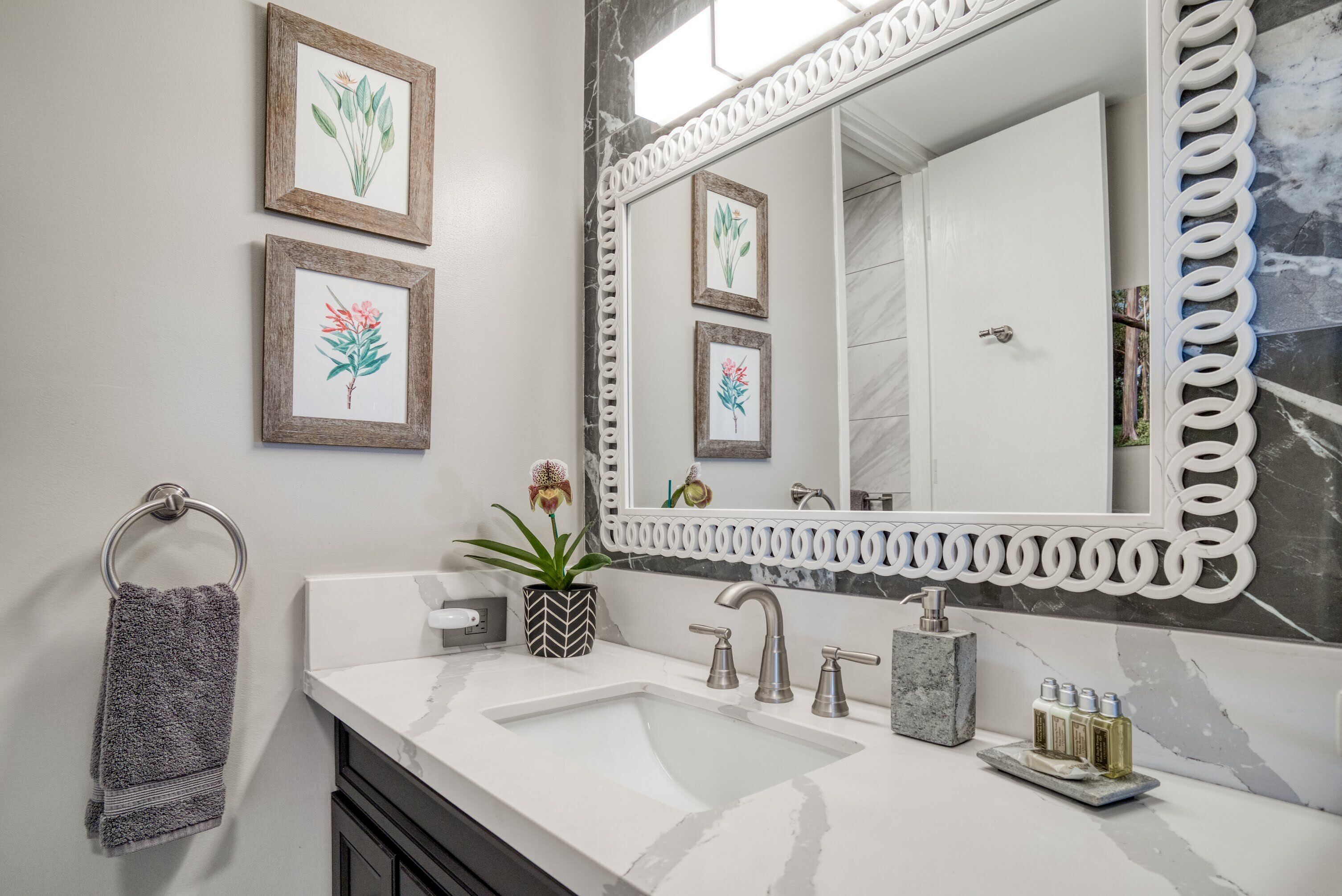 A bathroom with a white sink, two faucets, a large mirror with a decorative frame, and two framed botanical prints on the wall.