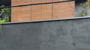 Fachada de edificio con revestimiento gris oscuro, una puerta de madera con tela morada y una pequeña ventana con marco metálico.