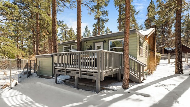 A cabin with a wooden deck and a metal railing, surrounded by tall pine trees and covered in snow. - Bear's Booth (Big Bear City)