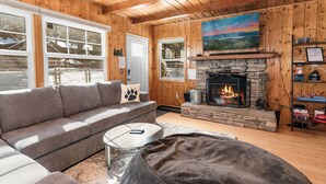 A cozy cabin interior with a stone fireplace, wooden walls, a gray sofa, and a large window overlooking a snowy landscape. - Bear's Booth (Big Bear City)