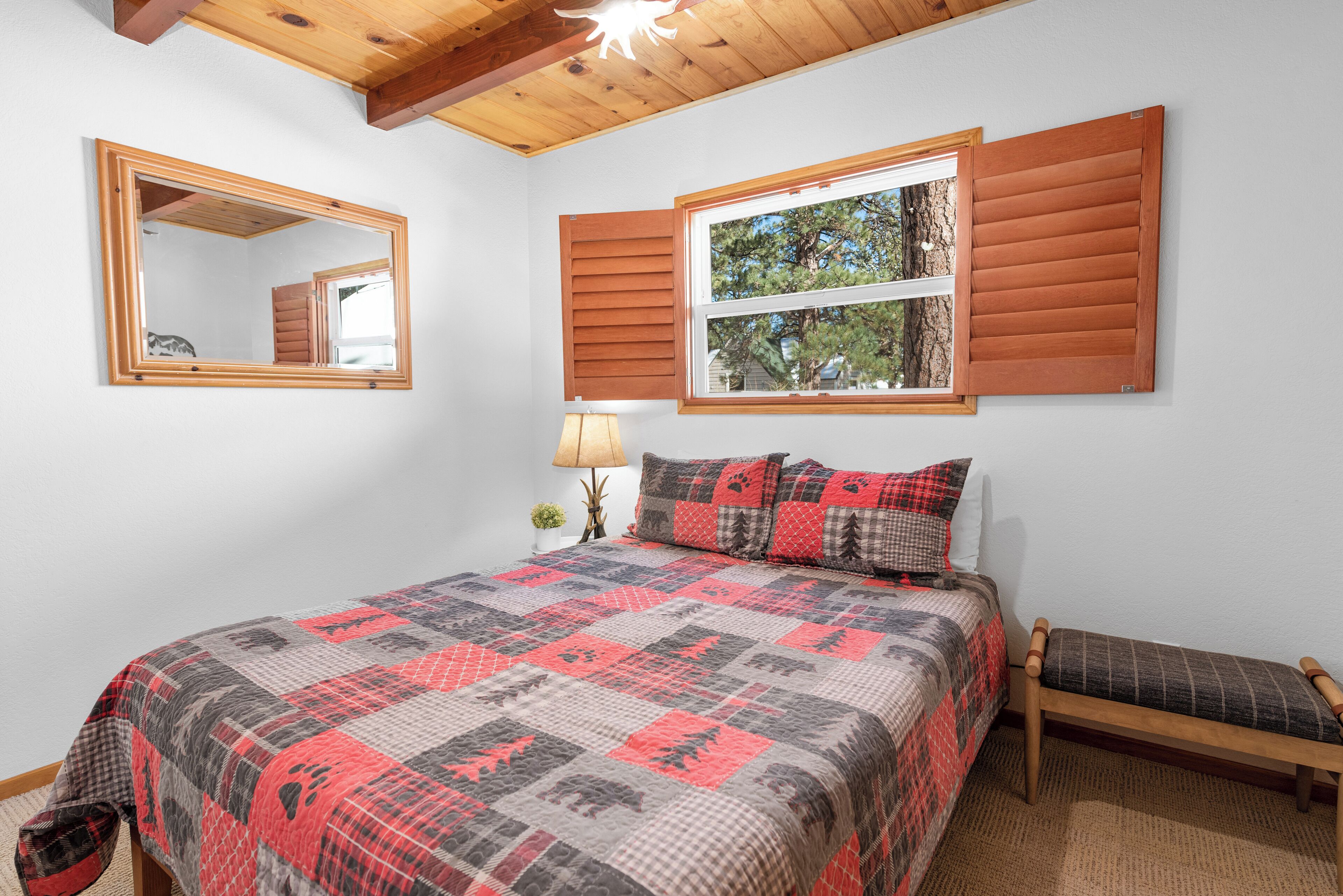 A bedroom with a large bed, a bedside table with a lamp, a bench, a window with shutters, and a mirror on the wall.