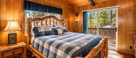 A wooden cabin bedroom with a bed, a nightstand, a lamp, and a window with a view of trees.
