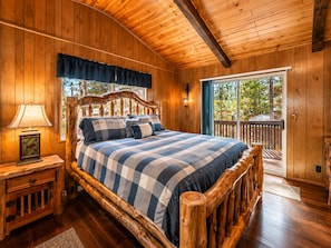 A wooden cabin bedroom with a bed, a nightstand, a lamp, and a window with a view of trees.