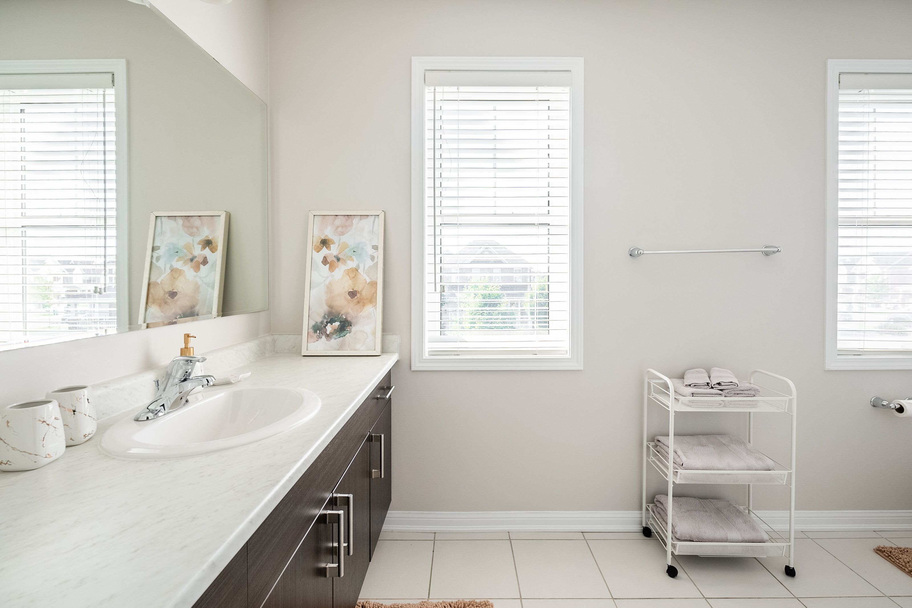 A bathroom with a sink, two windows with blinds, a towel rack, and a white shelf with towels.