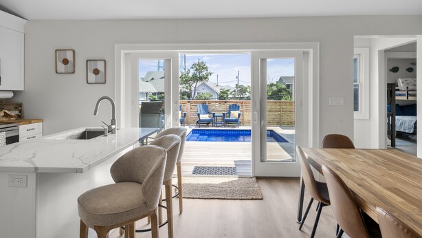 Une cuisine moderne dotée d’un grand îlot central, d’un coin repas avec une table et des chaises en bois, et offrant une vue sur un jardin avec une piscine et du mobilier d’extérieur.
