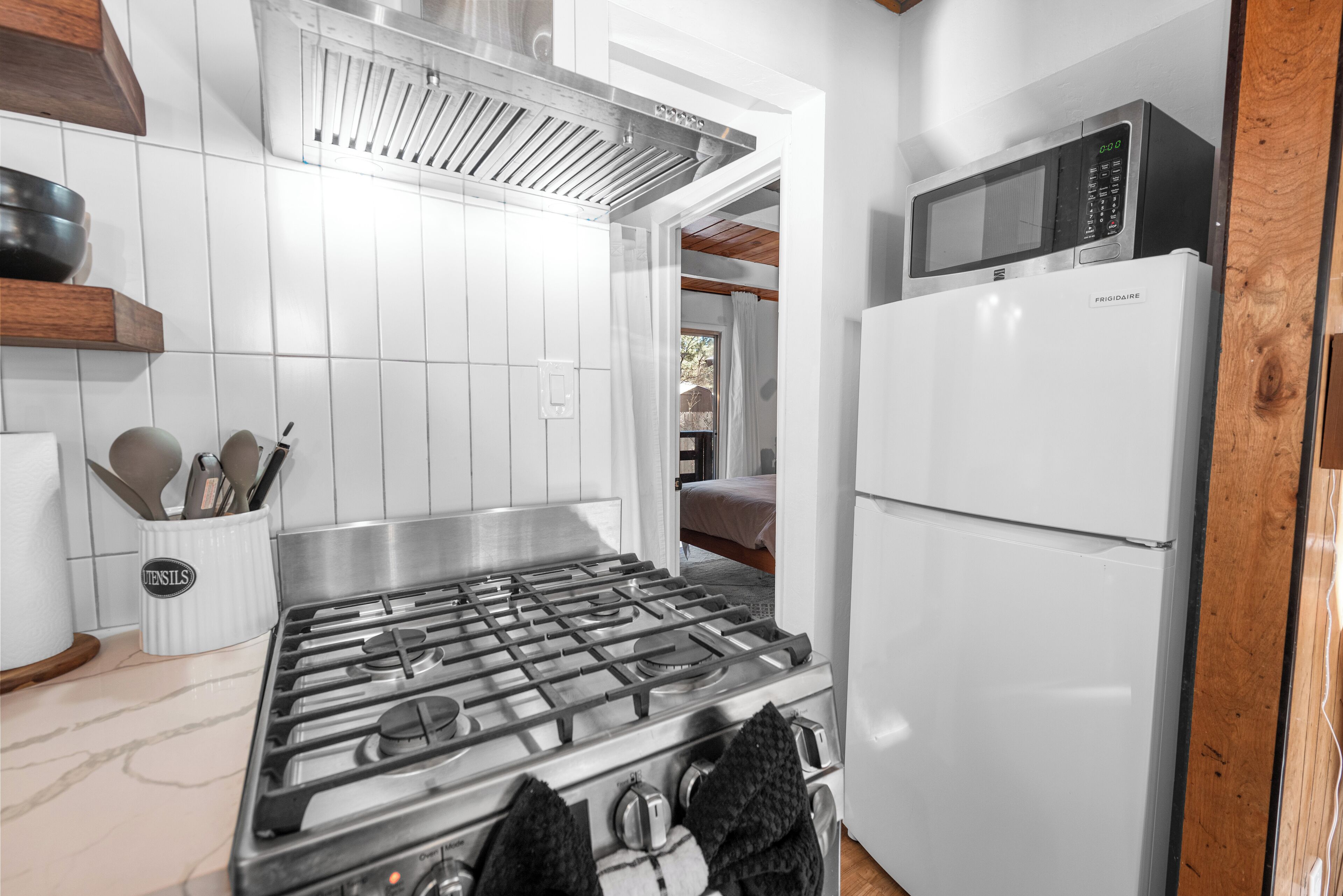 Una cocina con estufa a gas, microondas y refrigerador. Se observa un portal utensilios con herramientas de cocina y una ventana sobre la estufa.
