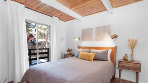 A bedroom with a large bed, wooden headboard, two bedside tables with lamps, and a view of a wooden deck outside. - Bearadise (Big Bear City)