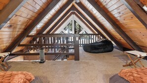 An attic bedroom with a sloped ceiling, wooden beams, a ceiling fan, a bed, a bean bag, and a view of the outdoors. - Bearadise (Big Bear City)