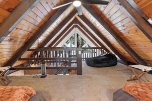 Un dormitorio en el ático con techo inclinado, vigas de madera, ventilador de techo, cama, sillón de mimbre y vistas al exterior.