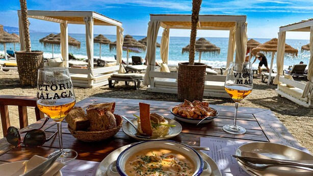 Un montaje para comer frente al mar, con una mesa preparada para dos personas, mostrando un tazón de sopa, un plato de pan y una copa de vino.