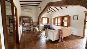 Un espacio habitable amplio con chimenea, sofá y televisor. Hay una estantería y una ventana con contraventanas.