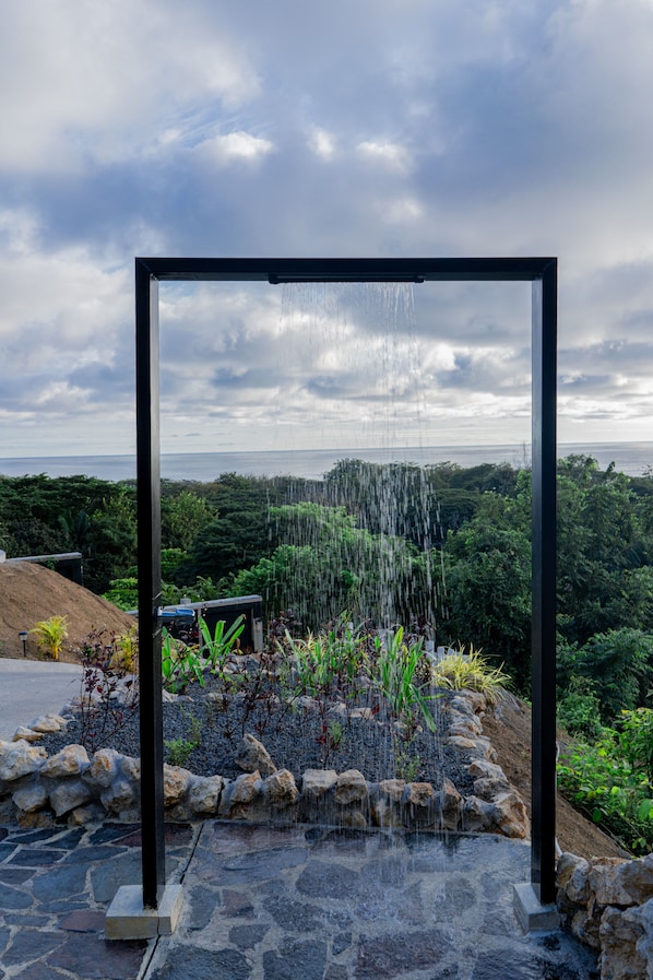 A modern outdoor water feature with a metal frame, surrounded by a stone base and lush greenery.