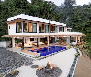 A modern two-story house with a large outdoor pool, a covered patio, and a fire pit.