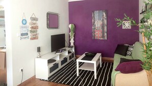 A living room with a white entertainment unit, a flat-screen TV, a small white table, and a green sofa with purple cushions. - Cozy small ground floor house for 4, 600 m from beach and close to city center (Costa da Caparica)