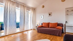 Un salón moderno con suelo de madera, un sofá con cojines naranjas, una mesa de centro blanca y vistas a través de grandes ventanas con cortinas estampadas.