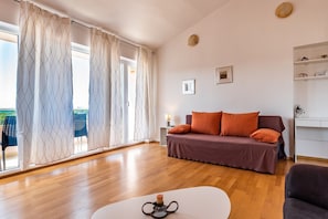 A modern living room with a wooden floor, a sofa with orange cushions, a white coffee table, and a view through large windows with patterned curtains.