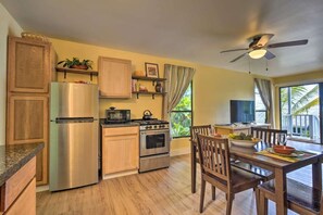 Una cocina compacta con refrigerador, estufa, microondas y gabinetes de madera. Hay una mesa de comedor con sillas y un televisor al fondo.