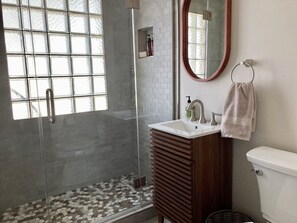 Un baño con ducha de vidrio, lavamanos blanco, tocador de madera, inodoro y espejo.