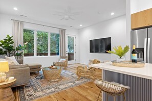 A modern living room with a sofa, coffee table, and chairs, a kitchen with a fridge and a TV, and large windows with curtains.