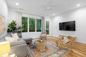 A spacious living room with a grey sofa, wicker chairs, a patterned rug, and a large window with curtains.