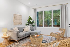 A modern living room with a grey sectional sofa, wicker chairs, a round coffee table, and a large window with curtains.