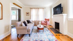 A living room with a beige sofa, a pink armchair, a coffee table, a fireplace, and a TV mounted above it. - Newly Restored 2BR Heights Cottage (Little Rock)