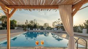 A pool area with a wooden pergola, two glasses of a beverage on a wooden stool, and lounge chairs with towels. - Aegean Apartments (Hersonissos)