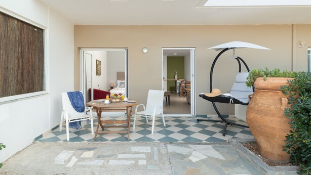 A patio area with a checkered floor, a table with chairs, a hanging chair, and a large terracotta pot with plants. - Aegean Apartments (Hersonissos)