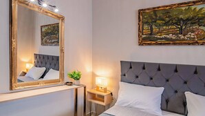 A bedroom with a large mirror, a painting, a tufted headboard, a bedside table with a lamp, and a bed with white bedding. - Napoleon Olive Home 2bd 1ba (Hersonissos)