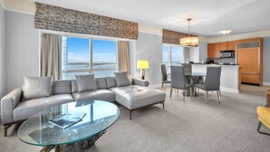 Ein modernes Hotelzimmer mit einer großen L-förmigen Couch, einem Glastisch, einem Essbereich mit einer kleinen Kochinsel und einer Küche mit Holzausstattung und einer Mikrowelle.