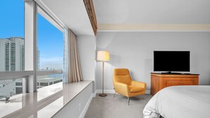 Ein Hotelzimmer mit großem Fenster, einem Bett, einem Fernseher, einem Sessel und einer Holzkommode.