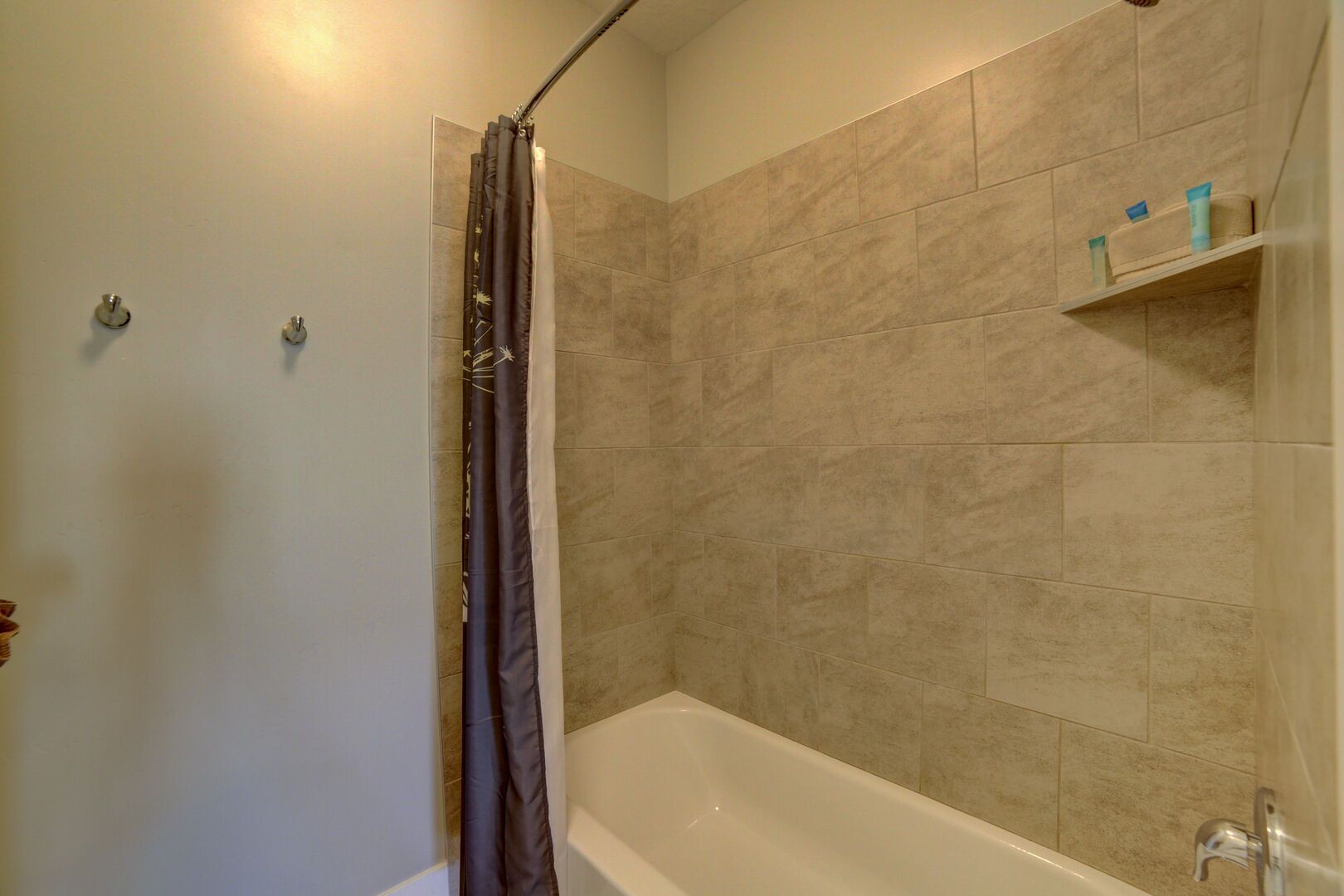 A bathroom with a bathtub, beige tiled walls, a shower curtain, and a shelf with toiletries.