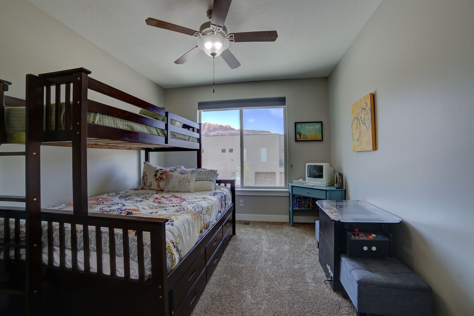Une chambre avec un lit superposé, un bureau avec un ordinateur, un ventilateur de plafond et une fenêtre donnant sur des immeubles.