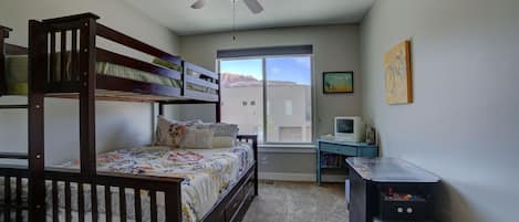 Une chambre avec un lit superposé, un bureau avec un ordinateur, un ventilateur de plafond et une fenêtre donnant sur des immeubles.