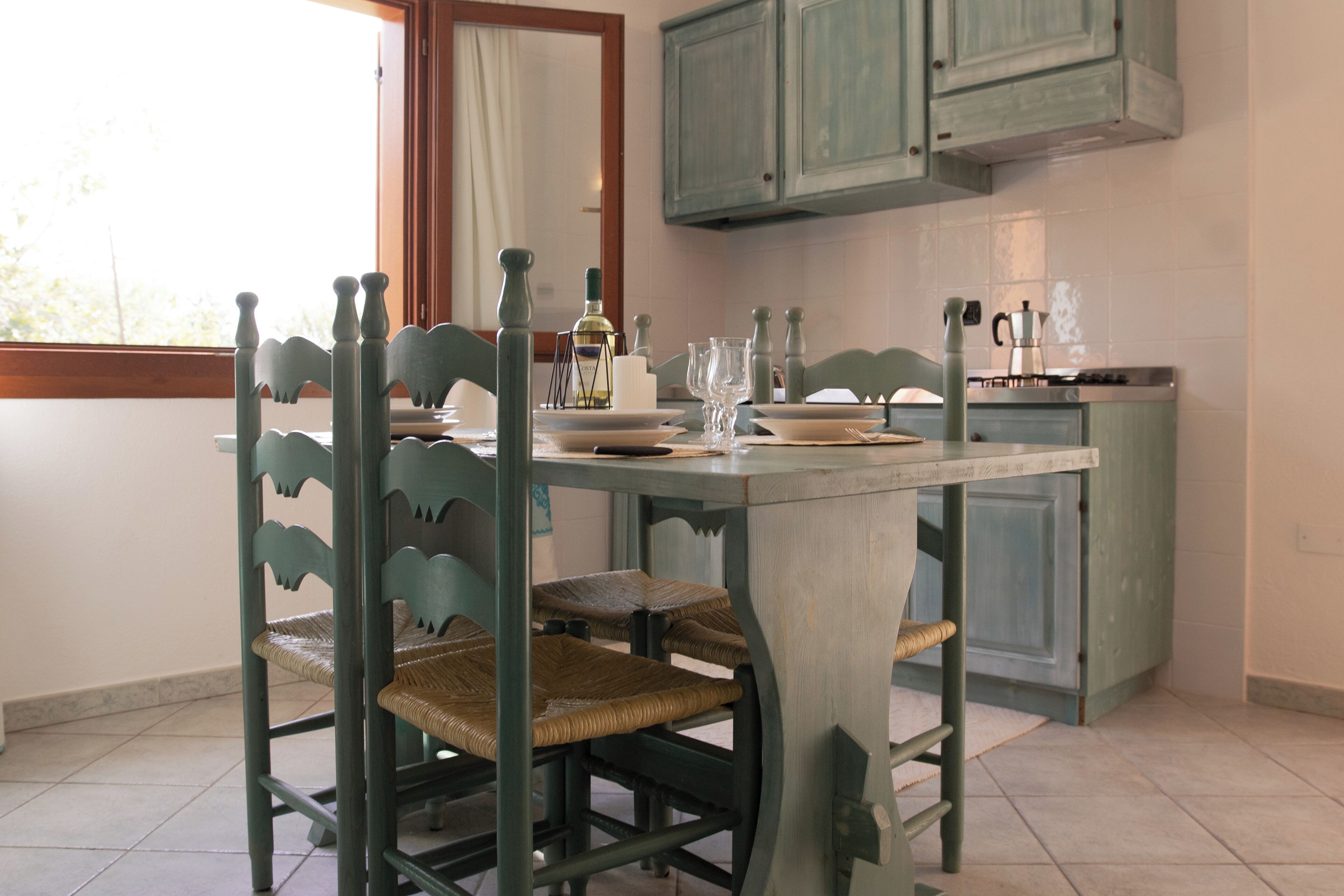 A dining area with a table set for four, surrounded by chairs with a unique design. A kitchen with teal cabinets is visible in the background.