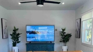 A living room with a brown leather sofa, a flat-screen TV mounted on a blue cabinet, a ceiling fan, and two potted plants. - Atlanta Home - 10 min from everything! (Atlanta)