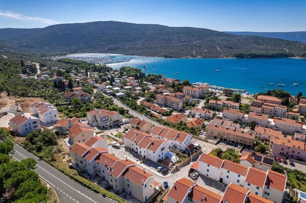 Une zone résidentielle côtière, avec des bâtiments aux toits de tuiles rouges, une mer d’un bleu limpide et un paysage vallonné en arrière-plan.