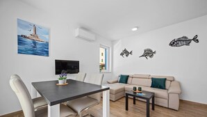 A modern living room with a dining area, a beige sofa, a TV, and wall art including a lighthouse painting and fish decals. - Apartment Tim (Cres)