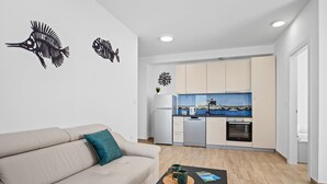 A modern living room with a beige sofa, a small coffee table with two mugs, and a view of a compact kitchen with a blue backsplash and a small island. - Apartment Tim (Cres)