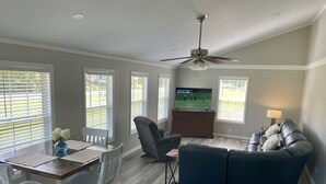 A living room with a dark blue sofa, a dining table with white chairs, a ceiling fan, and a television displaying a golf game.
