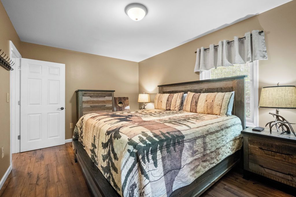 A bedroom with a bed, a nightstand, a lamp, a dresser, a door, and a window with curtains.