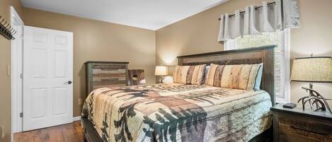 A bedroom with a bed, a nightstand, a lamp, a dresser, a door, and a window with curtains.