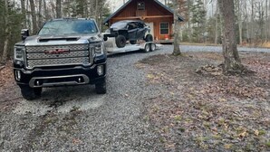 Ein schwarzer GMC-Truck steht auf einem Schotterweg in bewaldetem Gebiet, im Hintergrund ist eine kleine Hütte zu sehen.