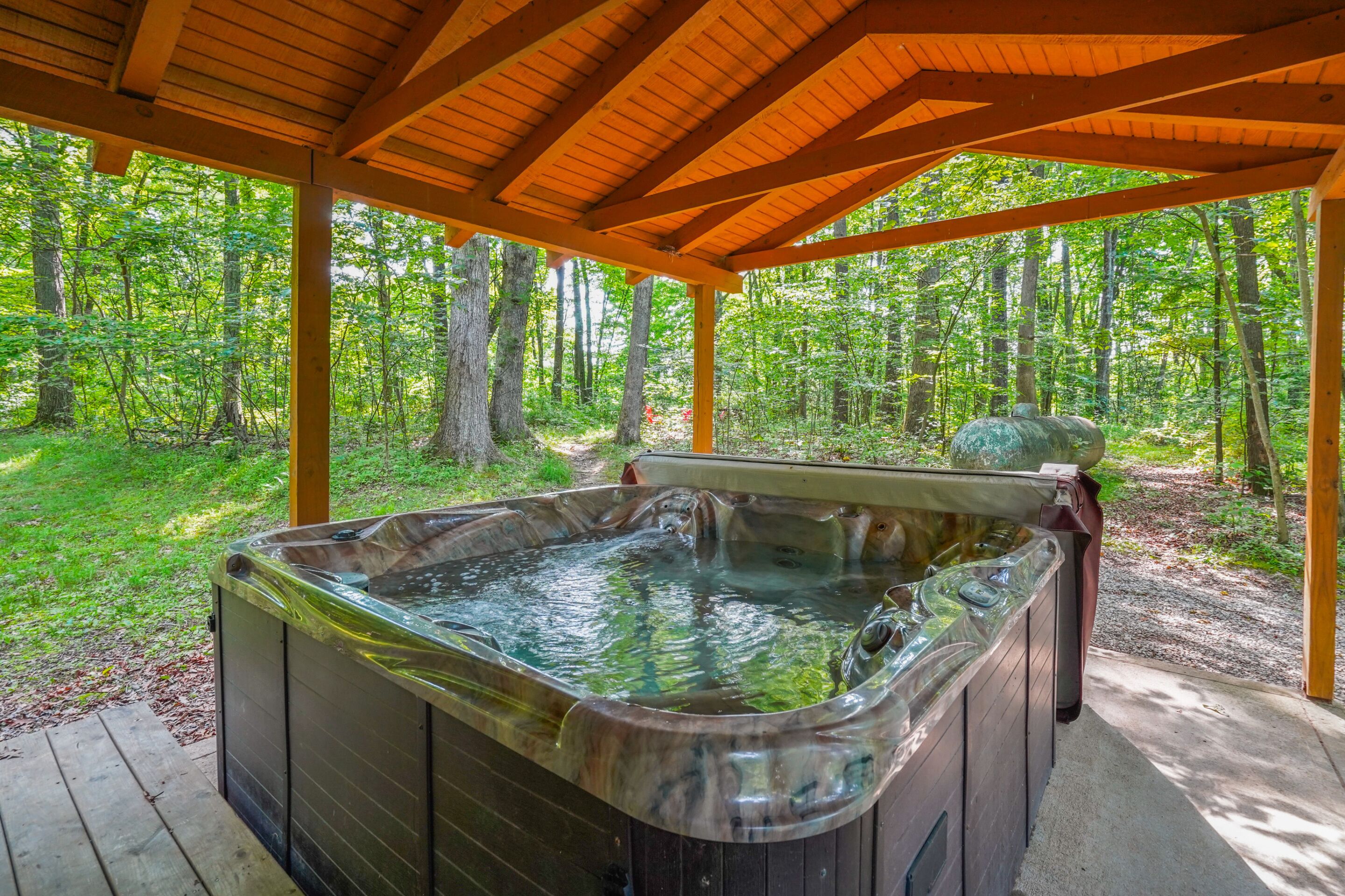 Un bain à remous installé sous un pavillon en bois, entouré d’une forêt.