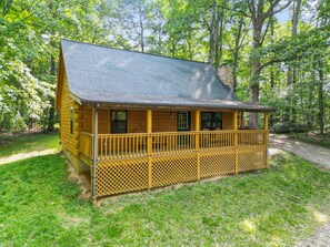 A wooden cabin with a porch and a chimney, surrounded by trees and a grassy area.