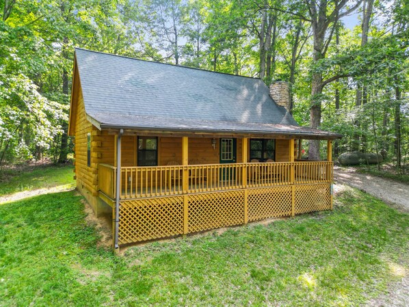 A wooden cabin with a porch and a chimney, surrounded by trees and a grassy area.